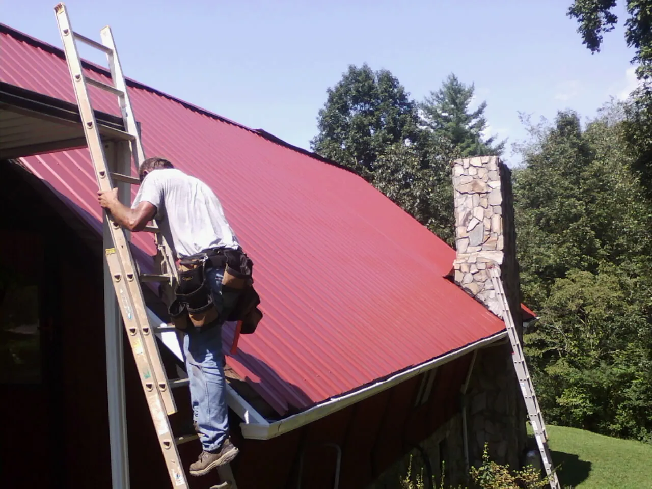 Roofer on a ladder beside a red metal roof and stone chimney — Soffit & Fascia Repair by Roofing Brothers in Commercial Point
